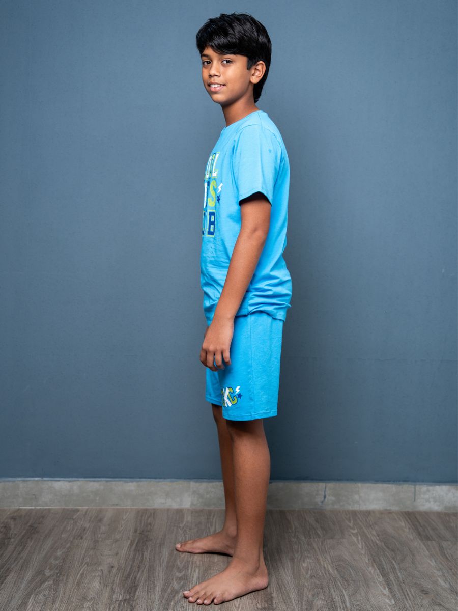 Young boy wearing a blue t-shirt and shorts against a gray wall.