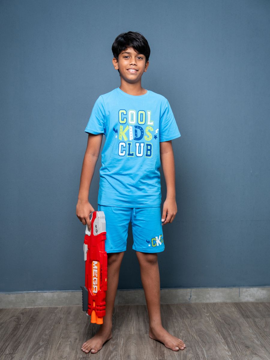Child wearing a blue ‘Cool Kids Club’ t-shirt and shorts, holding a toy gun against a gray background