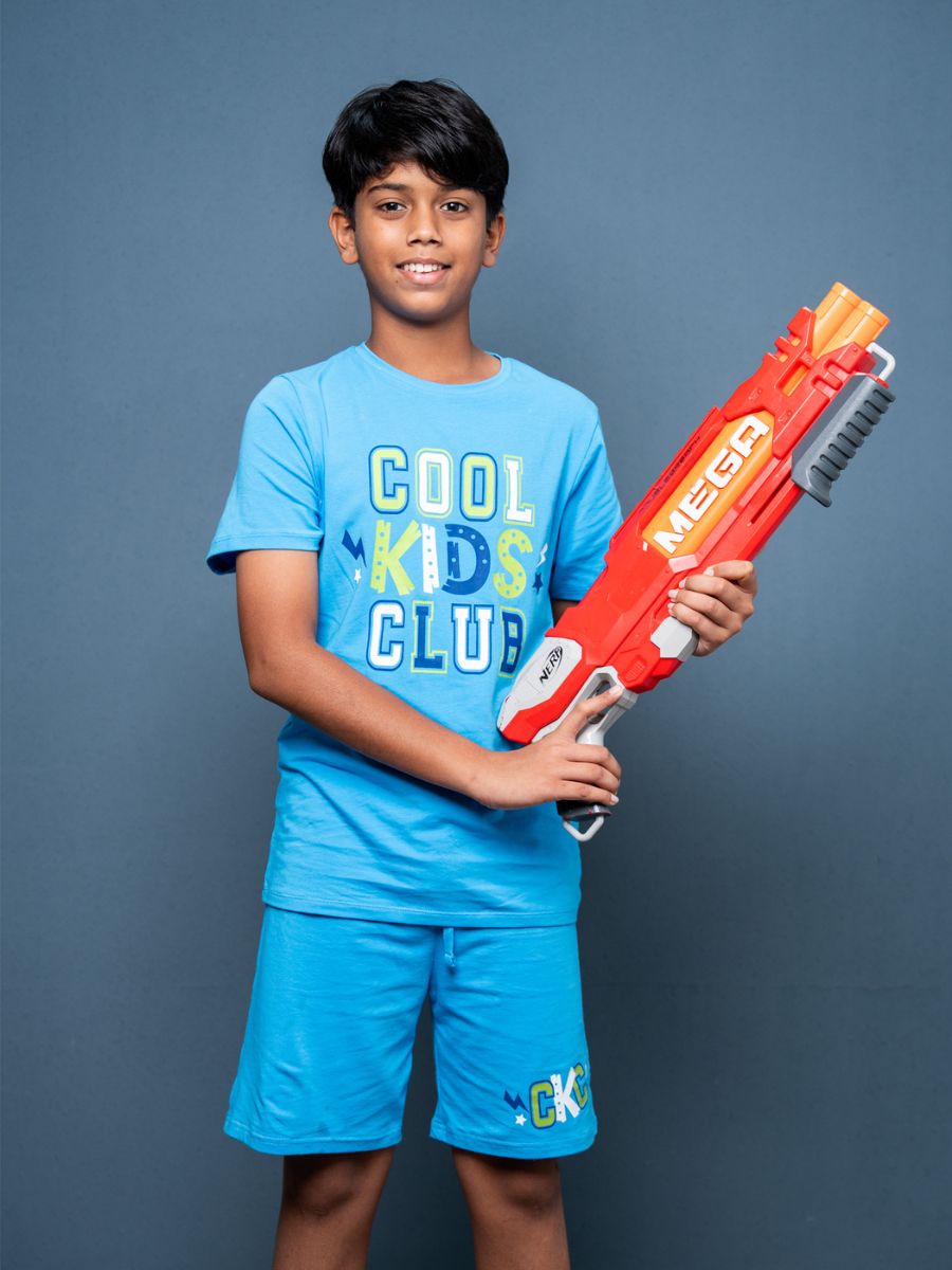 Child wearing a blue ‘Cool Kids Club’ shirt holding a red and white toy gun against a gray background