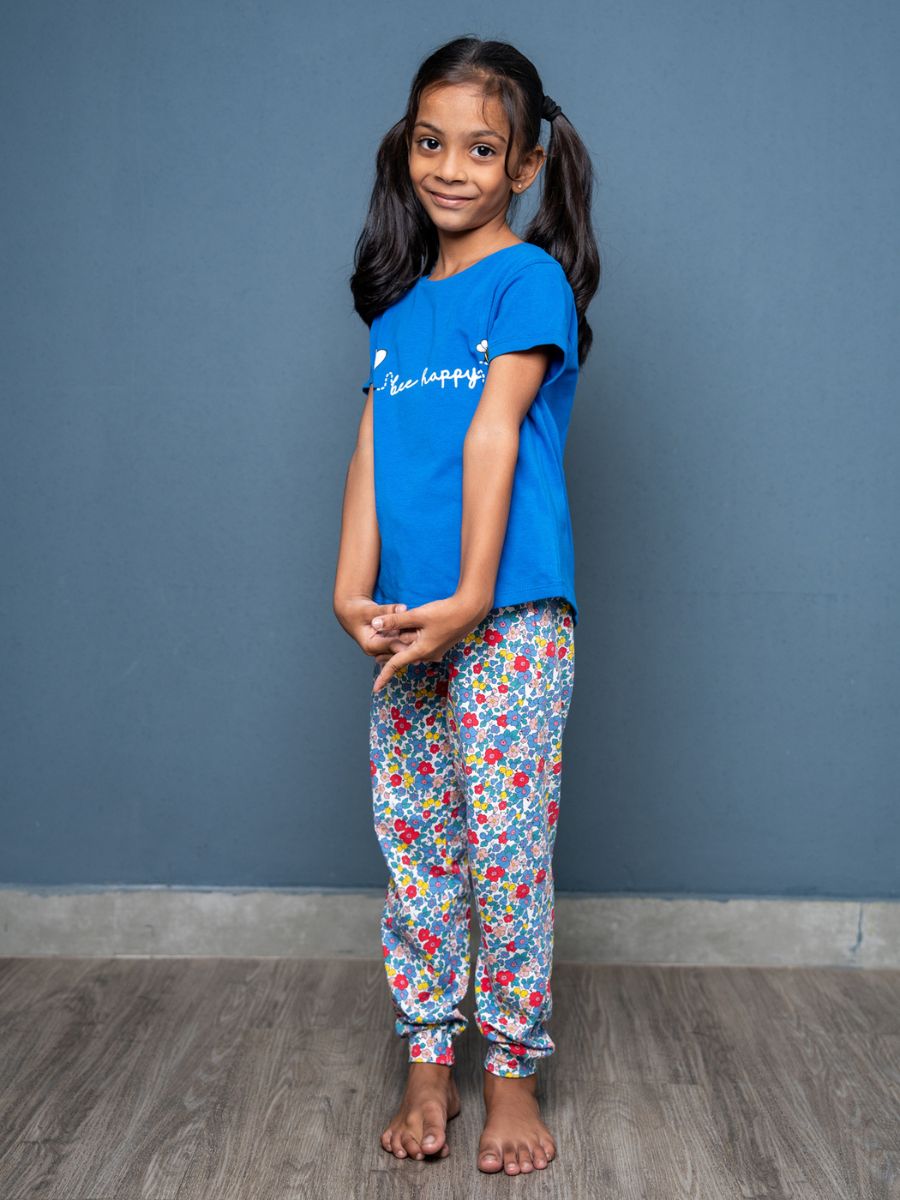 Young girl wearing a blue shirt and floral pants against a gray wall.