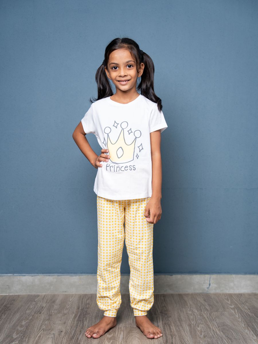 Young girl wearing a ‘Princess’ t-shirt and yellow checkered pants against a blue wall.