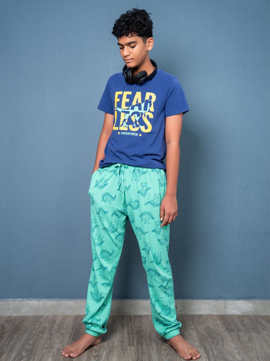 Person wearing a blue t-shirt with ‘FEAR LESS’ text and green patterned pants against a gray wall.