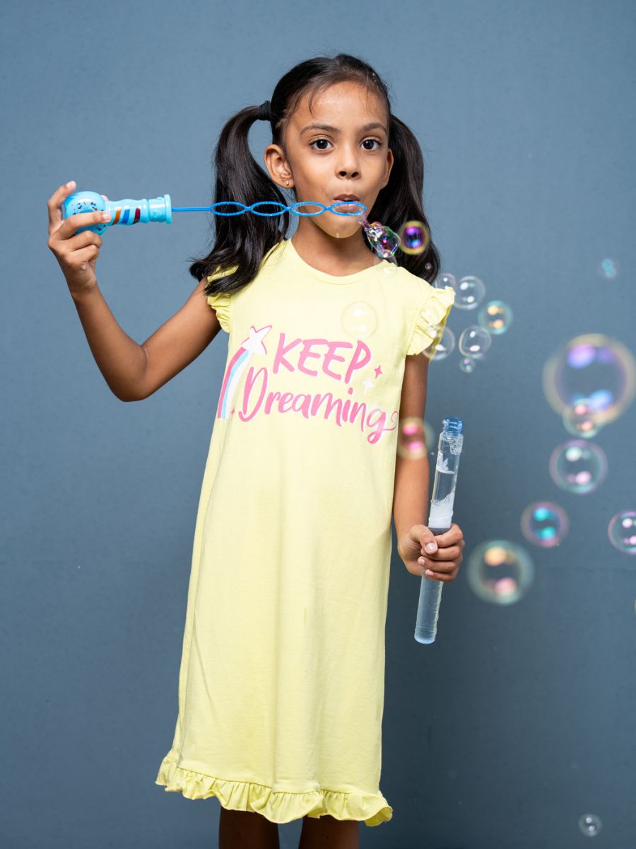 Girl blowing bubbles wearing a yellow nightgown with ‘Keep Dreaming’ text on a gray background