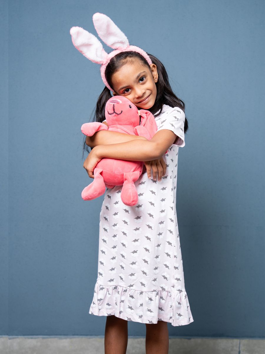 Young girl wearing a white dress with a pattern, holding a pink stuffed animal and bunny ears against a blue background